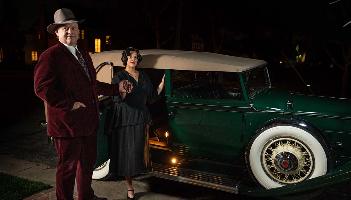 [image: A tall man in a dapper fedora and velour suit helps a stylish woman in a black dress out of a long green 1930s Packard automobile]