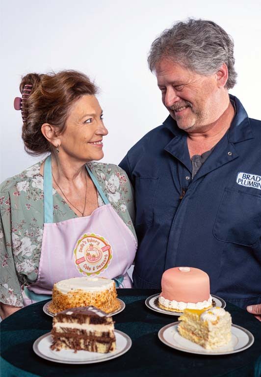 [image: A long-married couple, a white man and a white woman, look at each other lovingly behind a table of fancy cakes. She's wearing a "Della's Bakery" apron, and he's wearing a blue plumbing uniform.]
