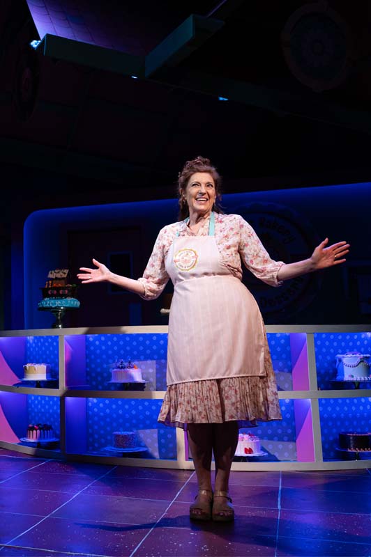 [image: A middle-aged white woman in a baker's apron and a frilly dress beams in the bright light of TV cameras]
