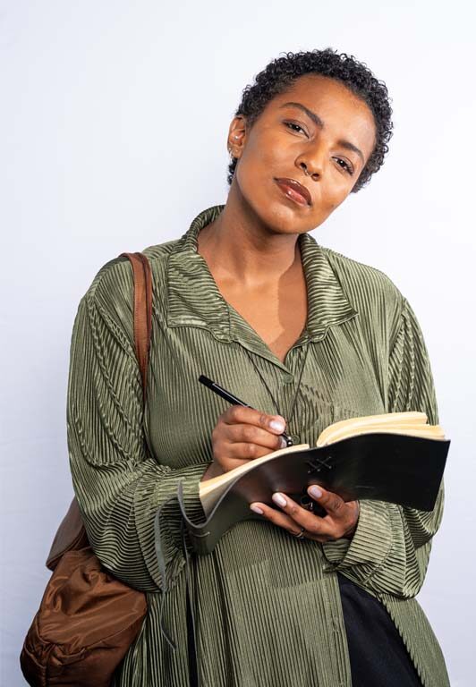 [image: A young Black woman with short hair holding a pen and notebook with a slightly skeptical writer's air]