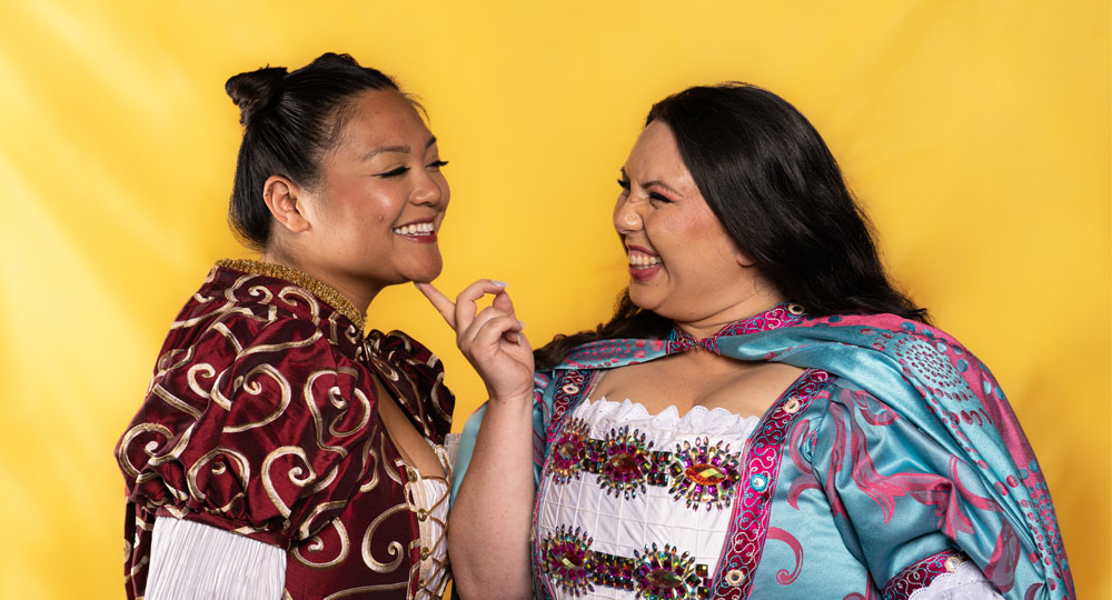 [image: Two female-presenting characters smiling into each other's eyes, one with her finger playfully under the other's chin. They wear opulent Elizabethan costumes.]