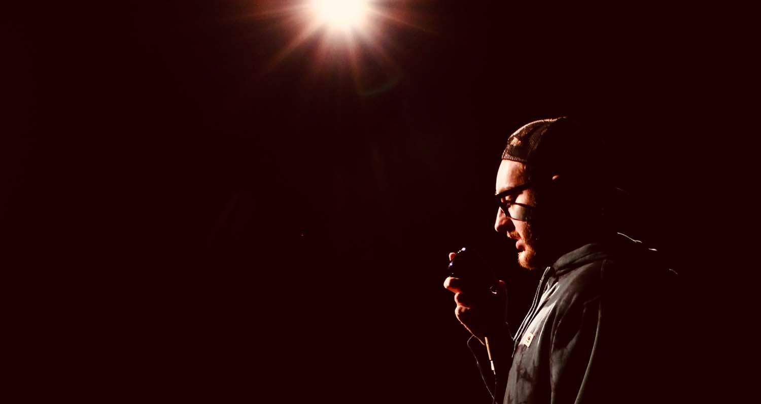 [image: A young white man in profile, holding a microphone, on a mostly darkened stage, with one bright light hitting him]