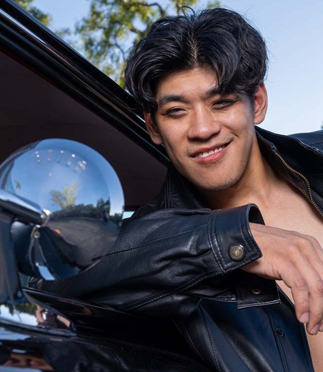 [Male actor with floppy black hair smiling seductively while leaning against a vintage black car]