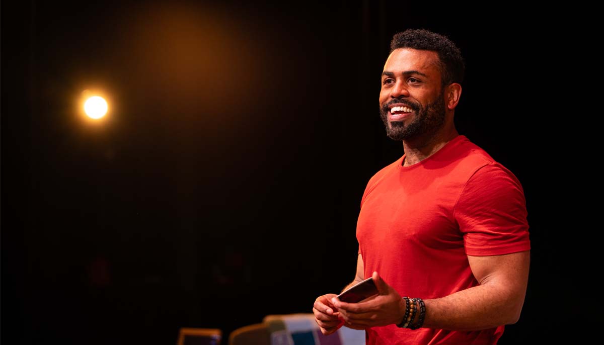 A smiling Black man holding his hands out in an open expression under a dramatic stage light.
