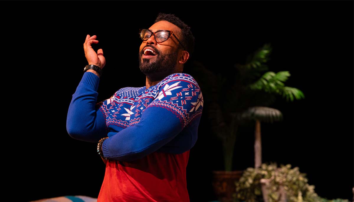 A smiling Black man playing the role of a fast-talking sales clerk, wearing cat-eye glasses and a too-small sweater.