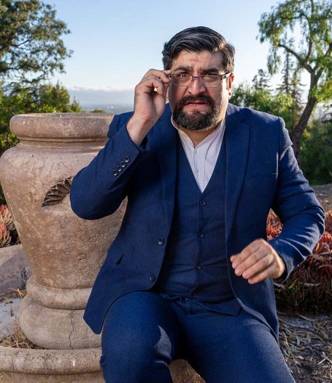 [Male actor with dark hair and goatee, in dark blue suit, looking confused while clutching his glasses]
