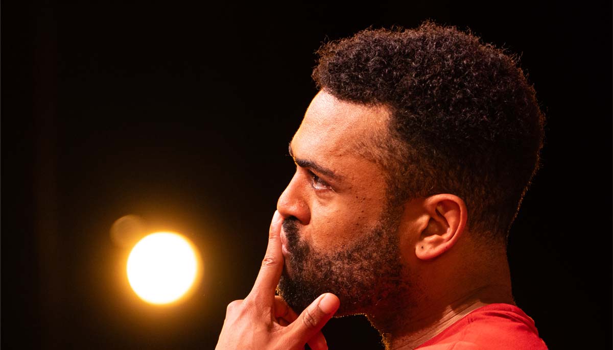 A young Black man looking pensive, a finger to his lips, viewed in profile under dramatic stage lighting.